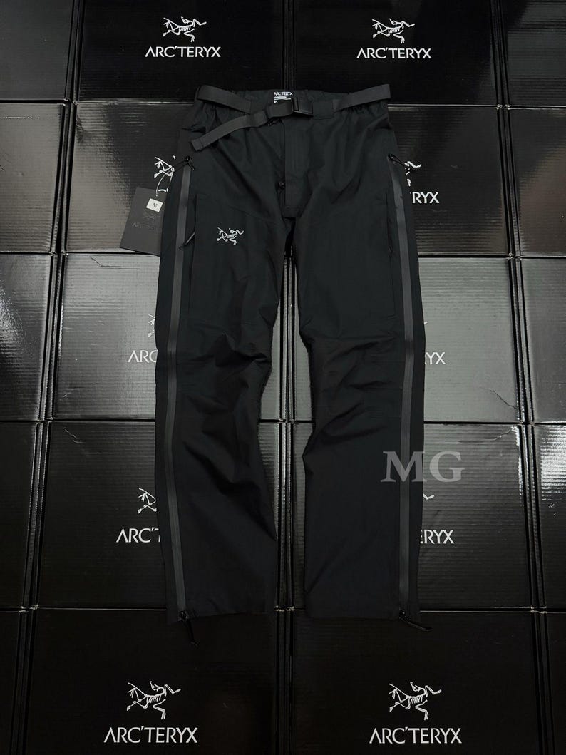 Puede incluir: Pantalones negros para actividades al aire libre con cintur&oacute;n, con el logotipo de Arc'teryx. Los pantalones tienen un dise&ntilde;o aerodin&aacute;mico con cremalleras y paneles reforzados. El fondo muestra cajas negras apiladas con el logotipo de Arc'teryx.