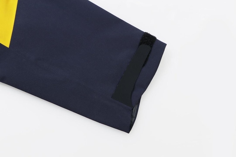 May include: Close-up of a navy blue garment sleeve with a yellow accent and a black Velcro closure. The fabric appears to be a smooth, possibly water-resistant material.