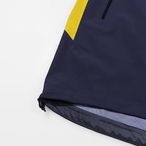 May include: Navy blue jacket with a yellow accent on the shoulder. The jacket has a black zipper and a drawstring at the bottom hem. The fabric appears to be water-resistant.