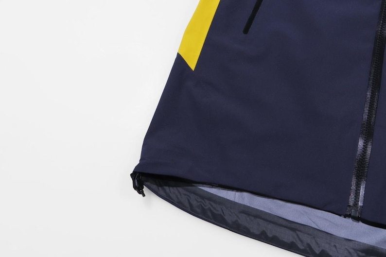 May include: Navy blue jacket with a yellow accent on the shoulder. The jacket has a black zipper and a drawstring at the bottom hem. The fabric appears to be water-resistant.