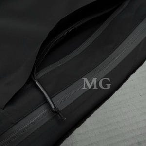 Puede incluir: Primer plano de una chaqueta negra con m&uacute;ltiples bolsillos con cremallera. La chaqueta presenta un dise&ntilde;o elegante y minimalista con las letras "MG" visibles en la tela. Las cremalleras son negras y el material parece ser una tela duradera y resistente a la intemperie.
