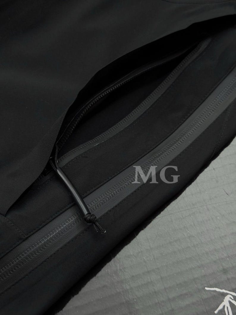 Puede incluir: Primer plano de una chaqueta negra con m&uacute;ltiples bolsillos con cremallera. La chaqueta presenta un dise&ntilde;o elegante y minimalista con las letras "MG" visibles en la tela. Las cremalleras son negras y el material parece ser una tela duradera y resistente a la intemperie.
