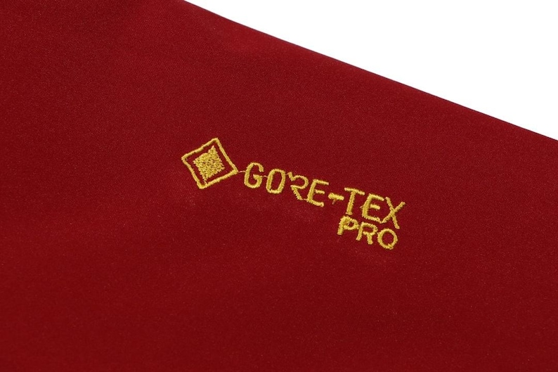 May include: Close-up of a section of red fabric with the embroidered text "GORE-TEX PRO" in gold. The fabric has a slightly textured appearance.