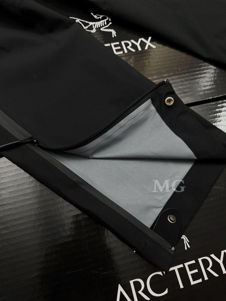 Puede incluir: Pantalones negros para exteriores con cremallera y forro interior gris. Los pantalones tienen un cierre a presi&oacute;n y un logotipo en la parte superior derecha. La marca "Arc'teryx" es visible en la parte inferior.