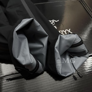 Puede incluir: Primer plano de una manga de chaqueta negra con forro interior gris. La manga tiene cierre de cremallera y un cord&oacute;n en el pu&ntilde;o. El logotipo de Arc'teryx es visible en el fondo.