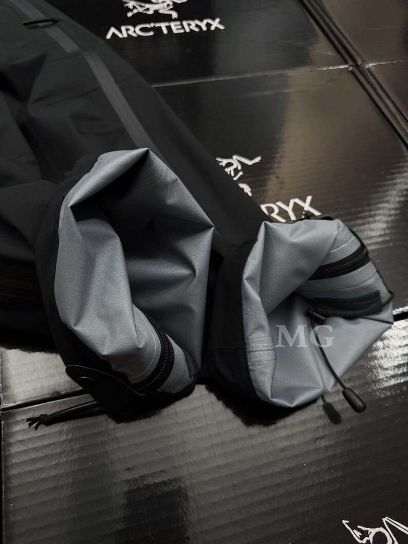 Puede incluir: Primer plano de una manga de chaqueta negra con forro interior gris. La manga tiene cierre de cremallera y un cord&oacute;n en el pu&ntilde;o. El logotipo de Arc'teryx es visible en el fondo.