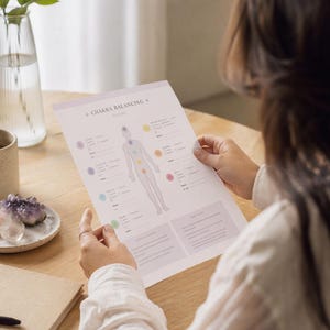 May include: A person holding a white paper with the text "Chakra Balancing" and a diagram of the human body with colored circles. A cup, a vase with greenery, and a small plate with crystals are on the wooden table.