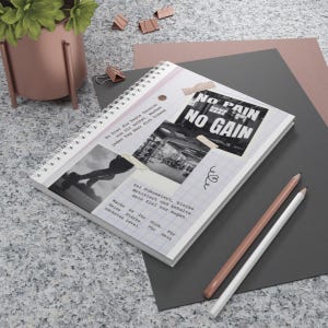 May include: A spiral-bound notebook with a white cover featuring motivational text and images. The notebook is open, displaying a collage of photos and handwritten notes. Two pencils, one white and one pink, lie beside the notebook.