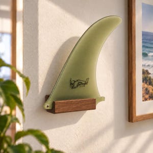 May include: A translucent sage green surfboard fin mounted on a rectangular dark wood base. The fin features a black graphic logo with the text Greenough Design 9.75-4A printed on its surface. The object is attached to a textured white wall.