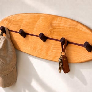 May include: A surfboard-shaped wall-mounted coat rack made of light-toned wood with a dark horizontal inlay stripe. It features five dark wooden pegs for hanging items. A tan baseball cap hangs on the left peg, and a set of keys hangs on the fourth.