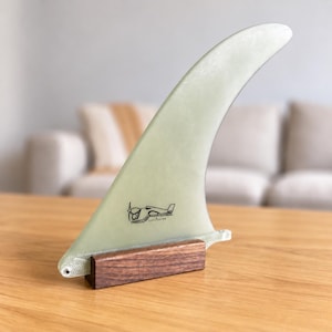 May include: A pale green, translucent surfboard fin with a curved, swept-back shape. The fin features a small black line-art graphic of a surfboard and is mounted upright in a rectangular, dark brown wooden base with a visible wood grain texture.