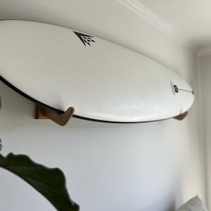 Peut inclure: Une planche de surf blanche avec des bordures noires et un logo graphique noir, montée horizontalement sur un mur à l'aide de deux supports muraux en bois. La planche présente un nez pointu et une forme allongée, maintenue par les supports.