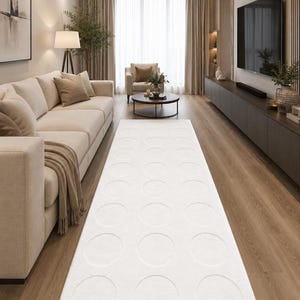 Könnte beinhalten: Ein modernes Wohnzimmer mit einem cremefarbenen Sofa, einem weißen Teppich mit kreisförmigen Mustern und einem großen Flachbildfernseher. Der Raum hat Holzböden und neutrale Vorhänge. Der Text "ojasvi décor" ist unten links sichtbar.