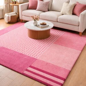 Könnte beinhalten: Ein rosa Teppich mit geometrischem Design. Der Teppich weist verschiedene Rosatöne auf, mit einem rechteckigen Rand und einem Mittelteil mit einem gepunkteten Muster. Der Teppich liegt auf einem Holzboden.
