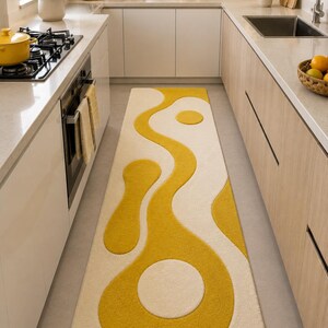 Könnte beinhalten: Ein langer, rechteckiger Teppich mit cremefarbenem Hintergrund und einem kräftigen, abstrakten Design in Senfgelb. Der Teppich weist fließende, organische Formen auf und schafft einen modernen Look. Der Teppich befindet sich in einer Küche.