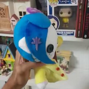 May include: A plush doll of a character from the movie "Coraline". The doll has a blue head, white face, and yellow outfit. A purple emblem is on the doll's face. The doll is held in front of a shelf with books and figurines.