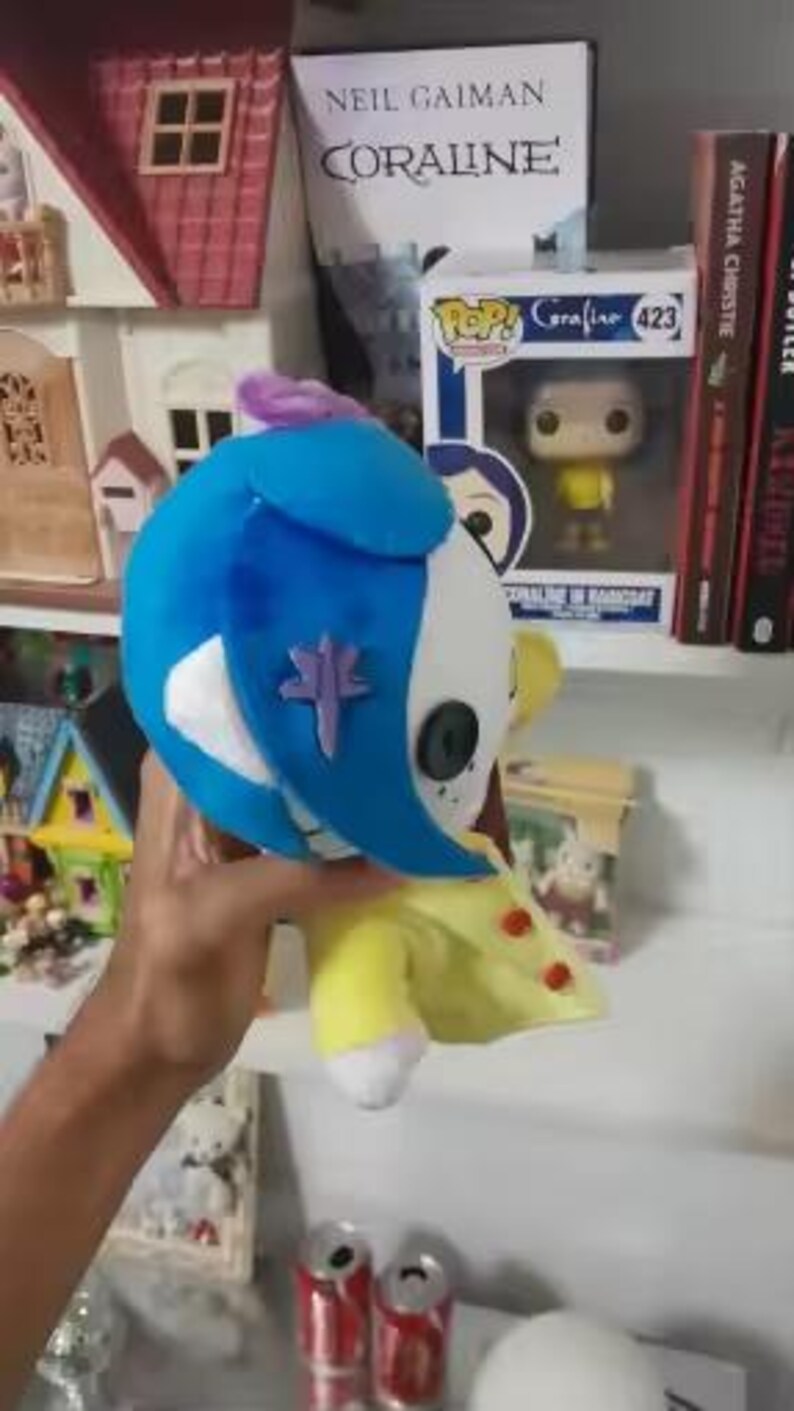 May include: A plush doll of a character from the movie "Coraline". The doll has a blue head, white face, and yellow outfit. A purple emblem is on the doll's face. The doll is held in front of a shelf with books and figurines.