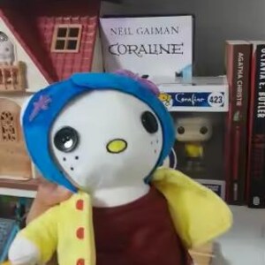 May include: A plush doll of a character from the book "Coraline". The doll has a white face with large black button eyes, a yellow oval mouth, and freckles. It wears a blue wig, a maroon shirt, a yellow jacket with red buttons, and blue shorts. The doll is held in front of a bookshelf.