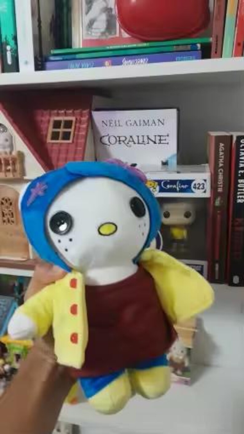 May include: A plush doll of a character from the book "Coraline". The doll has a white face with large black button eyes, a yellow oval mouth, and freckles. It wears a blue wig, a maroon shirt, a yellow jacket with red buttons, and blue shorts. The doll is held in front of a bookshelf.