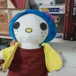 May include: A plush doll of a character from the book "Coraline". The doll has a white face with black button eyes, a blue wig, and a yellow jacket over a maroon shirt. The doll is holding its arms out.