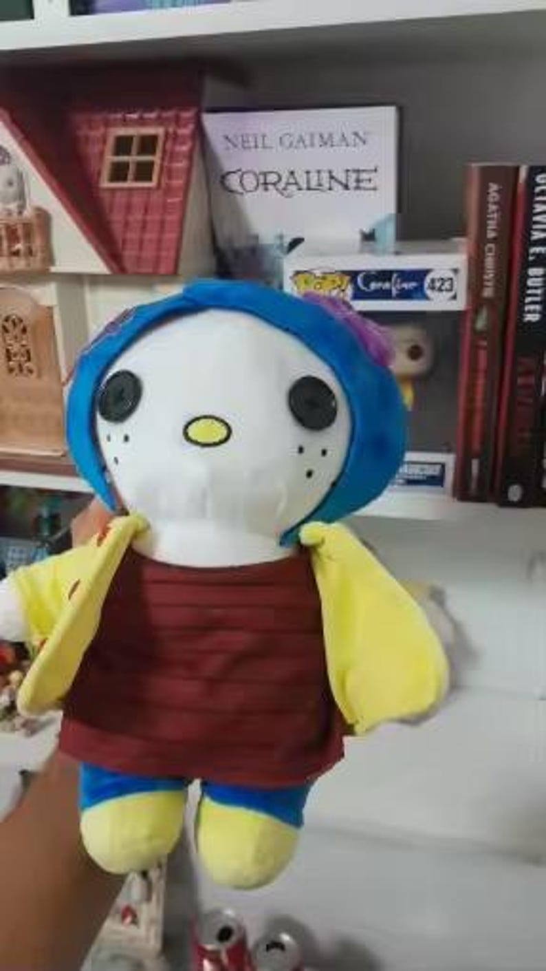 May include: A plush doll of a character from the book "Coraline". The doll has a white face with black button eyes, a blue wig, and a yellow jacket over a maroon shirt. The doll is holding its arms out.