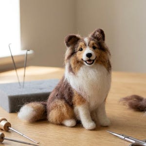 May include: A needle-felted Shetland Sheepdog figurine with brown, white, and black wool fibers. The dog is in a sitting position with a white chest and muzzle. Crafting tools including two felting needles, a foam block, and scissors are present.