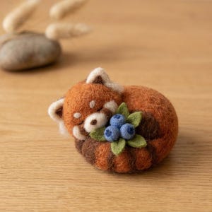 May include: A needle-felted red panda figurine curled into a ball, featuring rust-orange and dark brown fur, white facial markings, and a cluster of three small blue felted blueberries with green felt leaves tucked against its chest.