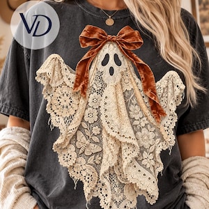 Pode incluir: Uma t-shirt cinzenta escura de manga curta com um gráfico de um fantasma feito de renda cor de creme. O design do fantasma é adornado com um laço de fita de veludo castanho no topo e inclui padrões intrincados de flores e traças.