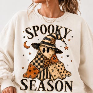 May include: A cream-coloured crewneck sweatshirt featuring a graphic of a ghost wearing a witch hat. The ghost is decorated with patchwork patterns including leopard print, checkers, and stars. The text SPOOKY SEASON is printed in black.