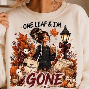 May include: A cream-colored sweatshirt featuring a graphic illustration of a person with a leopard print bow, holding a coffee cup. The design includes autumn leaves, pumpkins, ghosts, a street lamp, and the text ONE LEAF & I'M GONE.