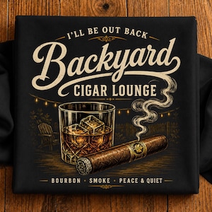 May include: A black t-shirt featuring a graphic with the text I'LL BE OUT BACK BACKYARD CIGAR LOUNGE and BOURBON SMOKE PEACE & QUIET. The design includes a glass of amber liquid with ice cubes, a lit cigar with rising smoke, and a wooden chair.