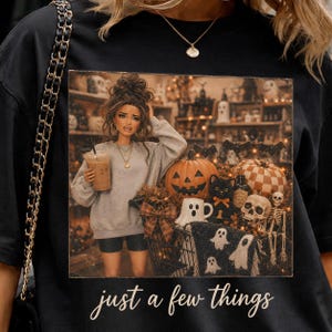 May include: A black t-shirt featuring a graphic print of a person in a gray sweatshirt holding an iced coffee, standing next to a shopping cart filled with Halloween items including a jack-o'-lantern, skull, and ghost mug, with the text just a few things.