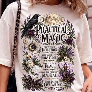 May include: A cream-colored t-shirt featuring a graphic print with a black raven, a full moon, purple flowers, a salt jar, and a moth. The text reads: Practical Magic, Trust Your Intuition, Keep Good Herbs Close, Protect Your Peace, Make The Ordinary Magical, Love Deeply, Live Wildly.