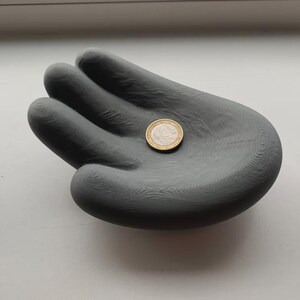 May include: A gray, hand-shaped decorative tray with a coin resting in the palm. The tray is made of a solid material and has a textured surface. The coin is gold and silver in color.