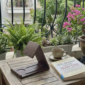 May include: A wooden phone stand with a dark brown finish sits on a wooden table. A cup of coffee and a book titled "The Power of Concentration" are also on the table, with plants and a balcony in the background.