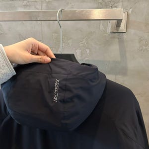 May include: A navy blue hooded jacket with the brand name "ARC'TERYX" in white lettering on the hood. The jacket is hanging on a metal hanger.