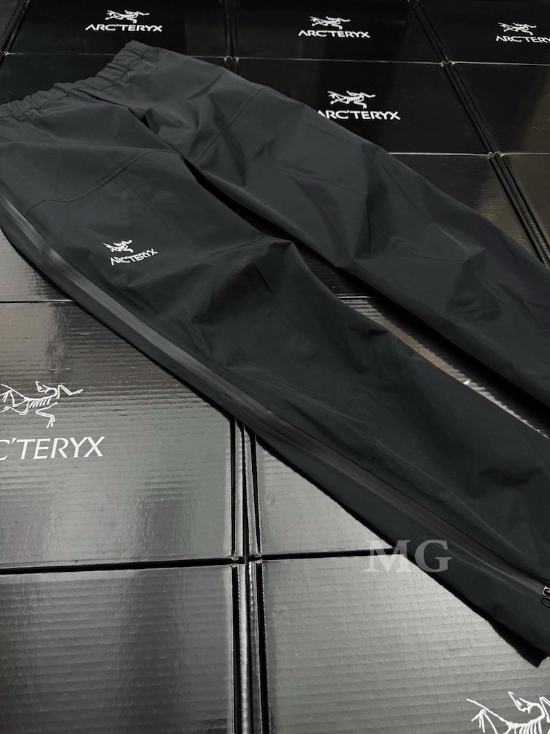 May include: Black waterproof pants with the Arc'teryx logo in white. The pants have a drawstring waist, a zippered leg opening, and a reflective stripe down the side. The pants are laid out on top of black boxes with the Arc'teryx logo.