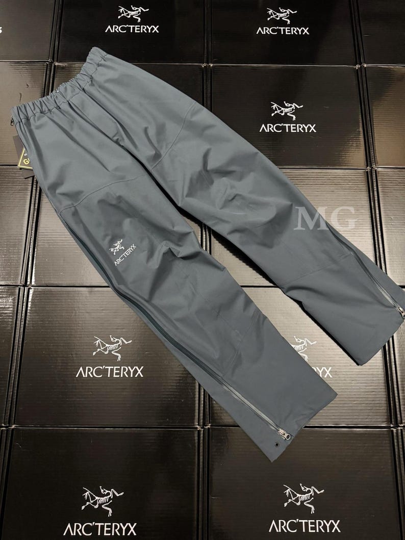 May include: Gray Arc'teryx rain pants with a drawstring waist and zippered pockets. The pants have the Arc'teryx logo on the left leg and zippers at the ankles. The pants are made of a lightweight, waterproof material.