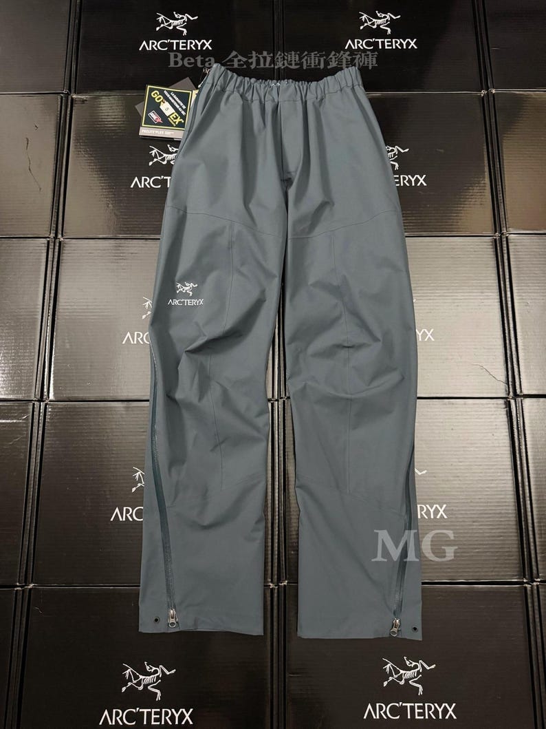 May include: Gray waterproof pants with the Arc'teryx logo on the leg. The pants have a drawstring waist and zippers along the sides of the legs. The pants are made of a durable, waterproof material.