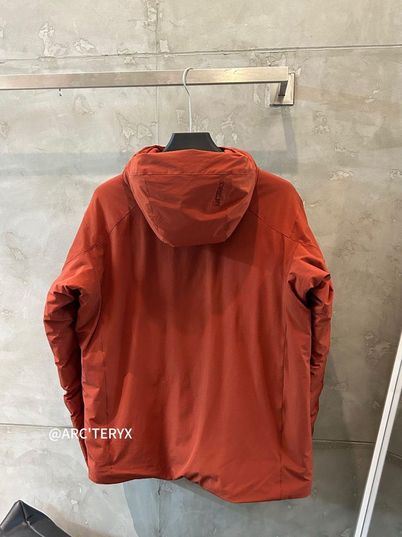 May include: Rust-colored hooded jacket hanging on a metal rack. The jacket has a full zipper and a hood. The brand name "ARC'TERYX" is visible on the hood and near the bottom of the jacket. The jacket is made of a lightweight material.