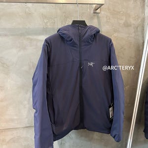 May include: A navy blue hooded jacket with a front zipper and the brand logo on the chest. The jacket is hanging on a metal rack against a gray wall. The text "@ARC'TERYX" is visible.
