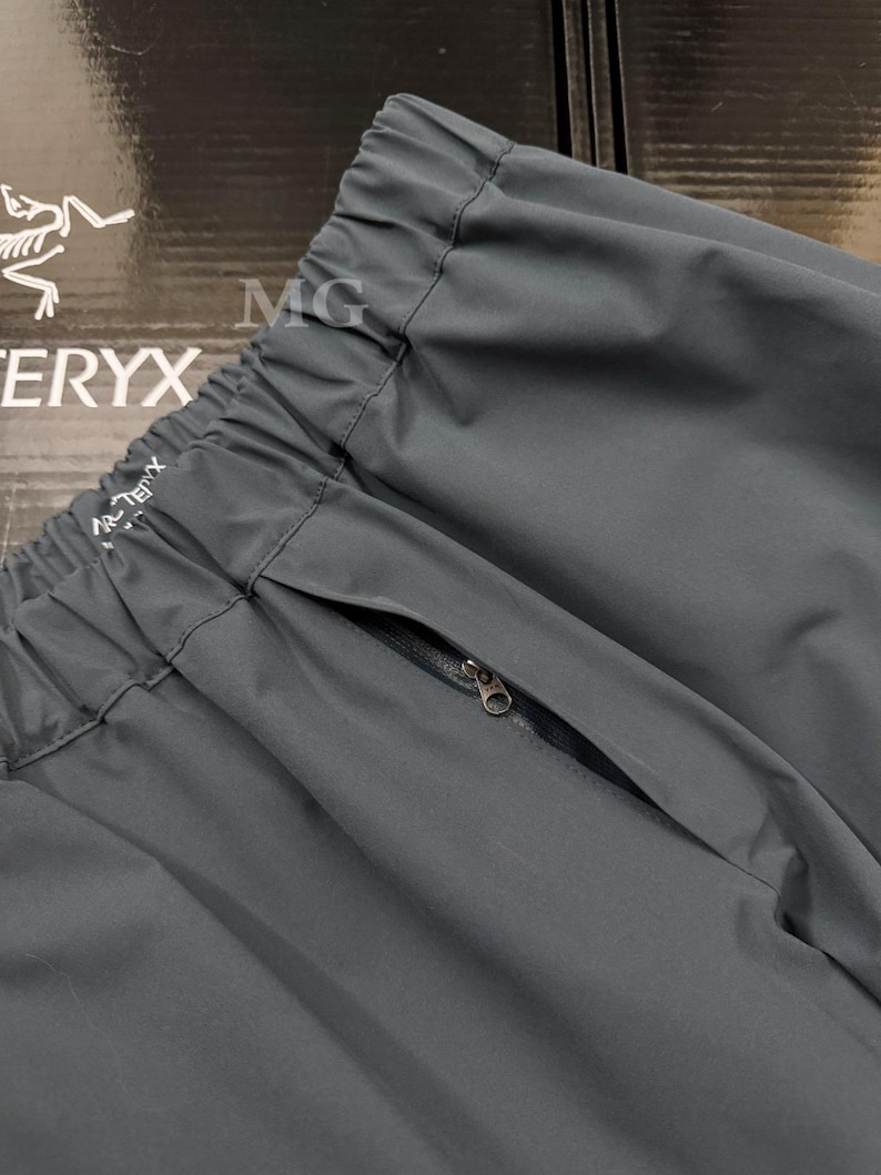 May include: Close-up of dark gray athletic pants with a zippered pocket. The pants have an elastic waistband and the brand name "Arc'teryx" is visible. The fabric appears to be a lightweight, durable material.