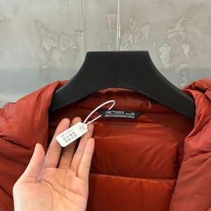 May include: A rust-colored Arc'teryx down jacket is displayed on a black hanger. The jacket has a quilted design and a tag attached with Chinese characters. The brand label is visible. The jacket is made of a shiny, smooth material.
