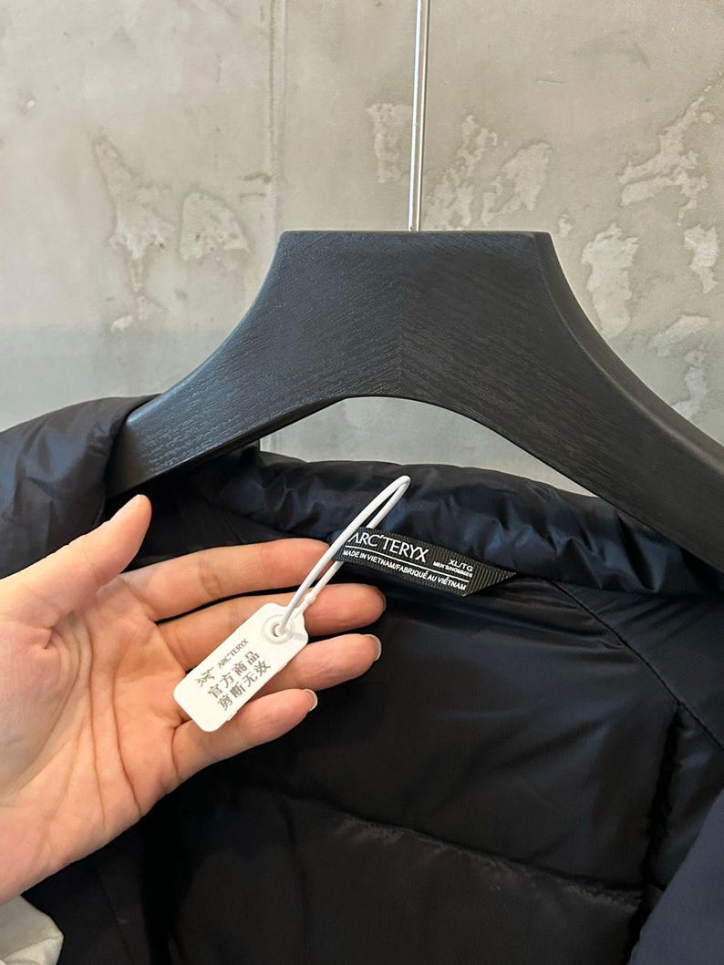 May include: Black puffer jacket with a tag attached. The tag has Chinese characters and the brand name. The jacket is hanging on a black hanger. The brand name is Arc'teryx.
