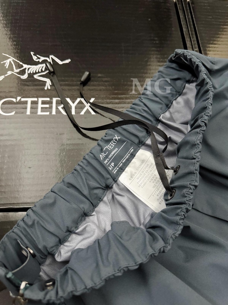 May include: Close-up of a pair of dark gray Arc'teryx pants. The pants have a drawstring waistband and a tag that reads "IN'S/HOMMES" and "S/P". The Arc'teryx logo is visible in the background. The pants are made in China.