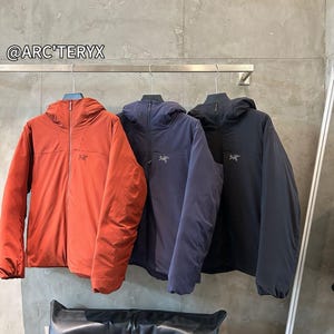May include: Three hooded jackets in rust, navy, and black hang on a metal rack. Each jacket has a full zipper and a small logo on the chest. The jackets are displayed against a neutral gray wall. The brand name "@ARC'TERYX" is visible.