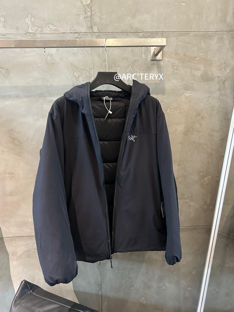 May include: A navy blue hooded jacket with a zipper closure, hanging on a black hanger. The jacket has a quilted interior and a small logo on the chest. The text "@ARC'TERYX" is visible.