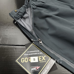 May include: Dark gray waterproof pants with a zipper detail and a tag that reads "GORE-TEX PRODUCTS". The pants have an elastic waistband and a small gold button. The tag also includes the text "LA GARANTIE DE VOUS MAINTENIR AU SEC".