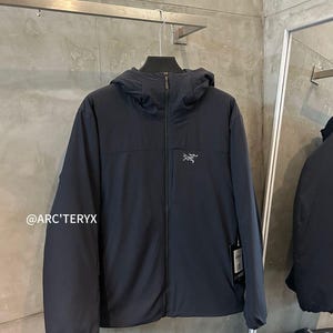 May include: A navy blue hooded jacket with a full-length zipper and the Arc'teryx logo on the chest. The jacket is hanging on a metal rack. The text "@ARC'TERYX" is visible in the lower left corner.
