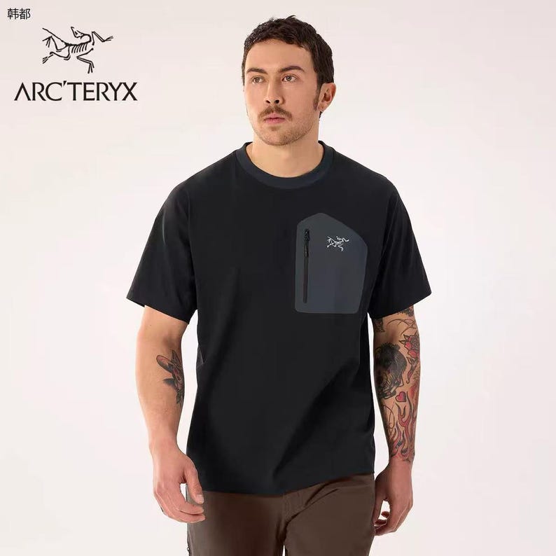 May include: Black short-sleeve t-shirt with a dark gray pocket on the chest. The pocket has a zipper and a small logo. The shirt has a crew neck with a dark blue trim. The brand name "ARC'TERYX" is printed on the shirt.